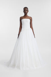 A model wearing an ivory strapless wedding gown with a structured asymmetrical corset and full tulle skirt, photographed against a seamless light grey studio background.