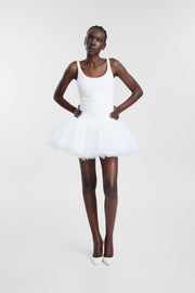 A model wearing a white mini tutu dress with a fitted scoop-neck bodice, thin shoulder straps and a voluminous tulle skirt, photographed against a seamless light grey studio background. 