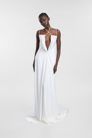 A model wearing an ivory satin wedding gown with a low cowl neckline, pearl strap detailing and a fluid floor-length skirt, photographed against a seamless light grey studio background.