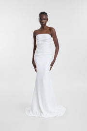A model wearing an ivory strapless wedding gown with horizontal draping, a fitted silhouette and floor-length skirt, photographed against a seamless light grey studio background.